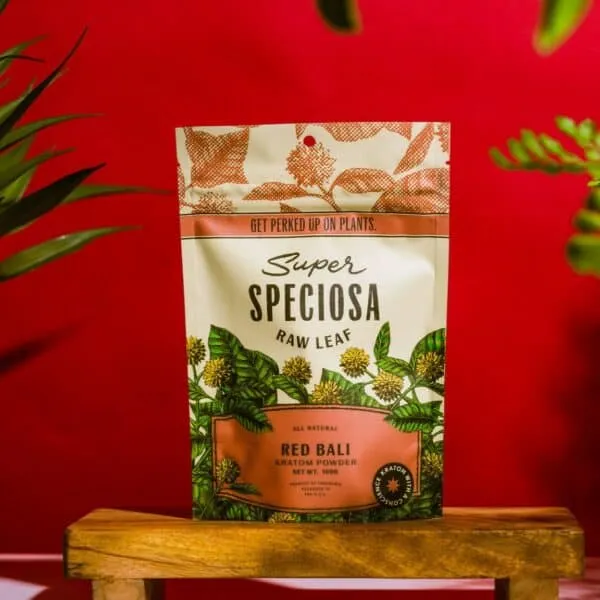 Red Bali Kratom Powder Bag displayed on wooden platform with plants and red background.
