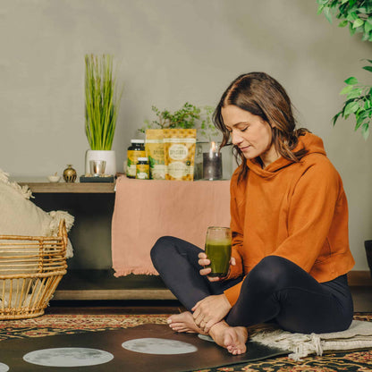 Woman is relaxing, drinking kratom drink, with assortment of Super Speciosa white kratom capsules and kratom powder products. White maeng da kratom bag in the background. Kratom products for body & mind shown.